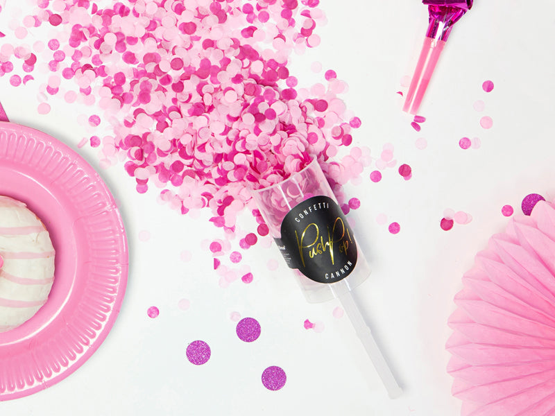 Roze Confetti Push Pop tussen uitgestrooide confetti, naast een roze bord, feesttoeter en papieren waaier.
