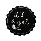 Zwarte ronde sticker met geschulpte rand, witte tekst ‘it's a girl’ en een klein bloemetje.