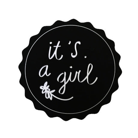 Zwarte ronde sticker met geschulpte rand, witte tekst ‘it's a girl’ en een klein bloemetje.