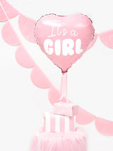 Roze hartvormige folieballon 'It's a Girl' boven roze cadeaudoosjes, met roze slingers op de achtergrond.