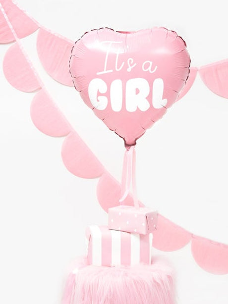Roze hartvormige folieballon 'It's a Girl' boven roze cadeaudoosjes, met roze slingers op de achtergrond.