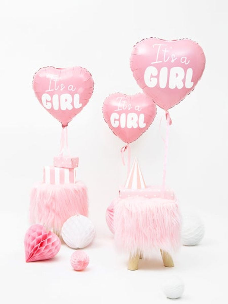 Drie roze hartvormige folieballonnen 'It's a Girl' bij roze krukjes, cadeaudoosjes en papieren decoraties.