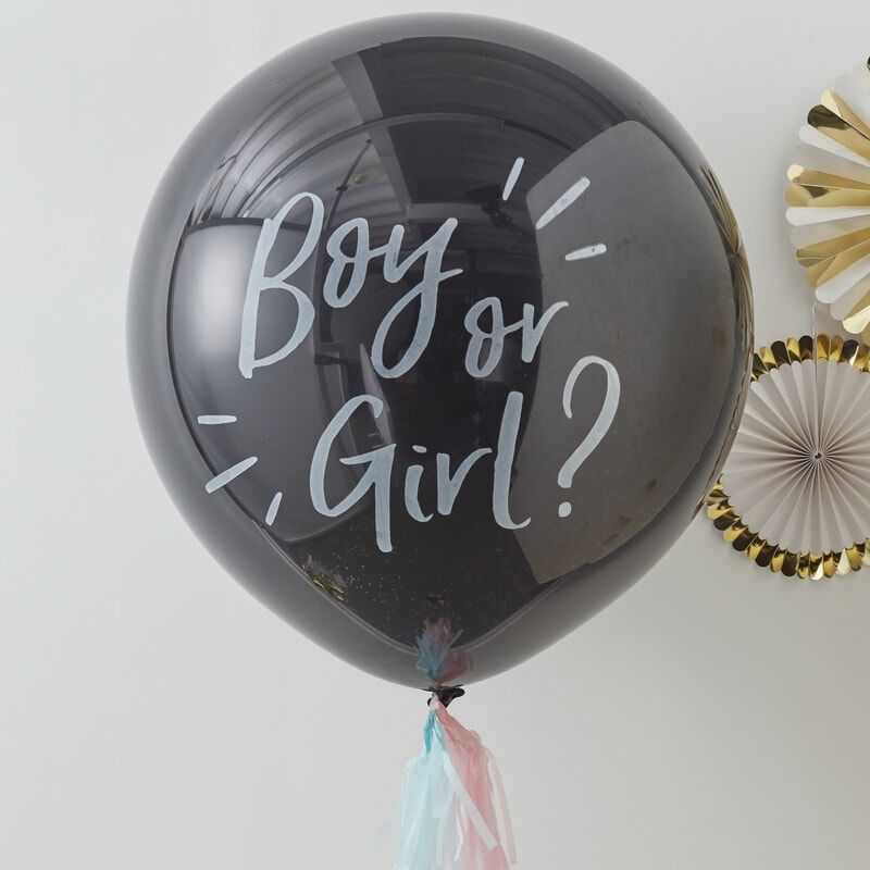 Zwarte gender reveal ballon met witte tekst ‘Boy or Girl?’, roze en blauwe kwastjes en papieren waaiers op de achtergrond
