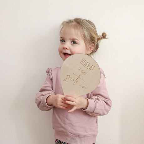Kind in roze kleding houdt een houten ballon met de tekst 'Hoera ik word grote zus' voor zich.