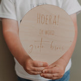 Kind houdt een lichtgekleurde houten ballon vast met de tekst 'Hoera! Ik word grote broer'.