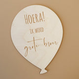 Lichtgekleurde houten ballon met de tekst 'Hoera! Ik word grote broer' op een bruine achtergrond.