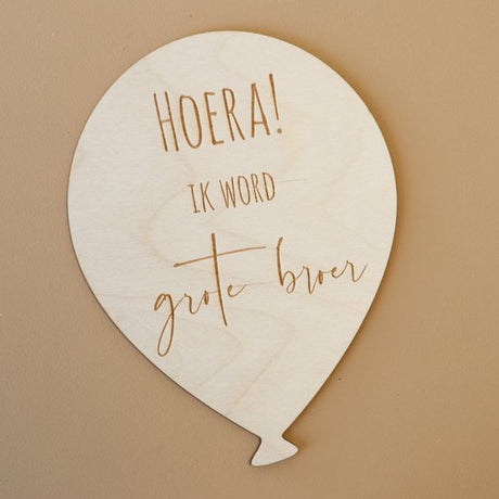 Lichtgekleurde houten ballon met de tekst 'Hoera! Ik word grote broer' op een bruine achtergrond.