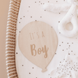 Houten ballon met opdruk ‘It's a boy’ op een gestippeld matras in een rieten mand, naast een witte knuffel.