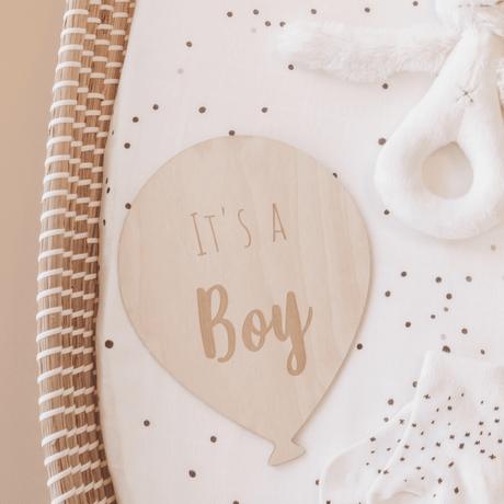 Houten ballon met opdruk ‘It's a boy’ op een gestippeld matras in een rieten mand, naast een witte knuffel.