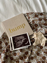 Bump zwangerschapsboek met nature kaft en gouden letters, naast echofoto's en babysokjes op een gebloemde ondergrond