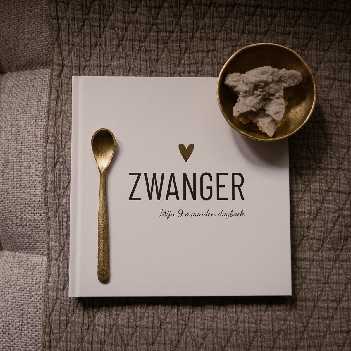 Wit zwangerschapsdagboek met goudkleurig hartje, lepel en schaaltje op grijze stof.