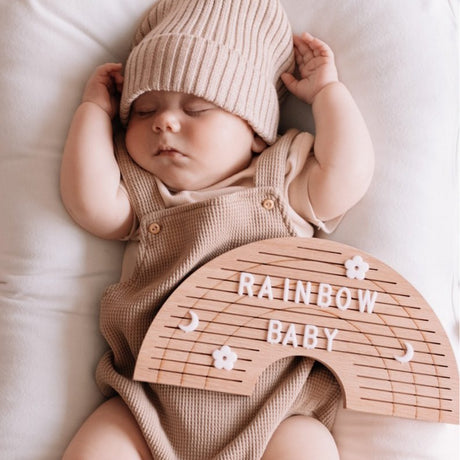 Houten regenboogletterbord met 'rainbow baby', bloemen en maantjes op de buik van een slapende baby.