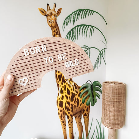 Houten regenboogletterbord met 'born to be wild' en een hartje, vastgehouden voor een giraf op de muur.