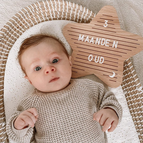 Houten letterbord in stervorm met witte tekst '3 maanden oud' naast een baby in een mand.