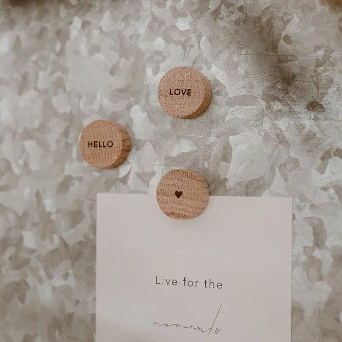 Drie ronde eikenhouten Hello Love-magneten met ‘Hello’, ‘Love’ en een hartje, waarvan één een kaart vasthoudt.