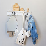 Witte magnetische kapstok met namen, een spiegel, tasjes en een spijkerjas, met houten decoraties op het plankje.