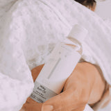 Hand houdt een fles Naïf Milky Badolie vast tegen een witte badjas met reliëf